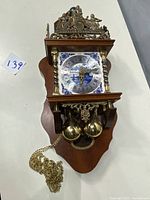 Full view of the vintage wooden wall clock with brass weights, decorated dial, and detailed brass top ornaments showing cherubs and figures.