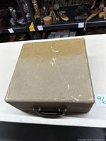 Closed textured beige carrying case with handle and latch for the typewriter, showing surface wear and scratches.