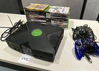 Full view of Xbox console, 23 stacked Xbox game cases, and two wired controllers on table.