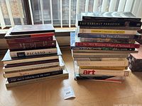 Two stacks of assorted art and cultural books arranged on a wooden parquet floor in front of a window with vertical blinds.