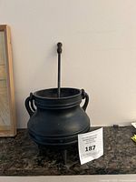 Full view of black cast iron cauldron with rounded body, handles, and tripod base, with pumice stone wand standing upright inside it.