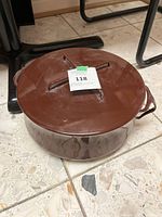 Round brown enamel pot with lid and two side handles on tiled floor, lot label on top