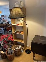 Full view of the black metal floor lamp standing beside shelving and a small table; the lamp shade is not included but a fabric shade is positioned over the bulb socket as a placeholder.
