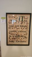 Framed vellum leaf showing Latin text and musical notes with illuminated initials, mounted on wall.