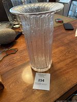 Vertical view of clear textured ribbed glass vase with flared rim and label with lot number 154 on wooden table.