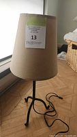 Full view of the table lamp showing the metal tripod base, cardboard shade, and electric cord on wooden floor near window.