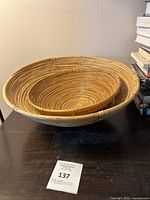 Two rattan woven bowls nested inside each other on a black surface with auction tag visible