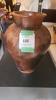 Front view of a large rounded brown clay vase with a wide neck and flared lip, placed on a black surface with an auction lot sticker marked 132.