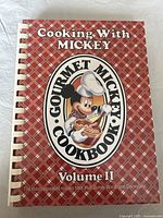 Front cover of the spiral bound cookbook Cooking With Mickey Gourmet Mickey Cookbook Volume II with red plaid design and Mickey Mouse chef illustration.