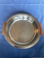 Top view of stainless steel serving dish on blue cloth showing round shape and wooden handles.