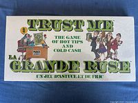 Front of the game box showing the title 'TRUST ME The Game of Hot Tips and Cold Cash' and colorful cartoon characters.