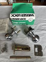 Photo showing Weiser Lock Passage Leverset with sleeve box, included latch assembly, screws, spindle and instructions laid out on a white sheet.