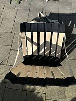 Photo showing one black and beige roll-up seat with canvas webbing laid flat on paved surface.