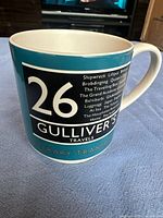 Front view of mug showing large '26 Gulliver's Travels' black and white label with literary locations around the black panel.