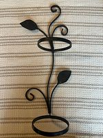 Front view of black wrought iron double plant pot holder placed on striped fabric, showing both 4-inch diameter rings and decorative curled metal work with leaves.