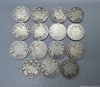Top down view of 15 Canadian silver quarters arranged in 3 rows of 5, reverse side facing up showing denomination and wreath design.