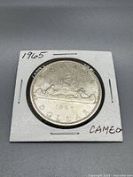Front side of 1965 Canadian silver dollar featuring the Voyageur design with depiction of canoe and Northern Lights, held in a white cardboard holder marked 1965 and CAMEO.