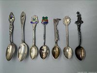 Top view of seven sterling silver souvenir spoons laid out side by side showing handle designs and enamel colors.