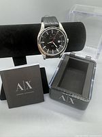 Photo of Armani Exchange wristwatch on display stand alongside its original case and brochure. Watch has silver stainless steel case, black dial, and black leather strap.