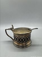 Side angle view of the sterling silver condiment pot with handle and hinged lid closed, showing detailed lattice pattern body and cobalt blue glass insert visible through the cutouts.