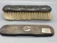 Front and back of a sterling silver topped grooming brush with floral design and natural bristles, showing detail of silver hallmark on underside.