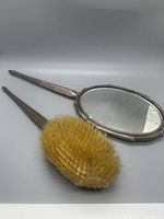 Photo showing the front of the hairbrush with natural bristles and the back of the hand mirror with smooth reflective surface and silver handle.