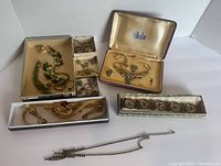 Photo showing an overview of various vintage costume jewellery pieces in boxes and loose including gold-tone mesh set with red stones, green rhinestone pieces, silver-tone pieces with blue stones in a velvet box, and a bracelet in a decorative box.