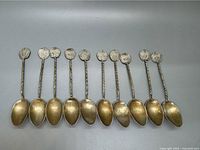 Top view of 10 silver tea spoons laid side by side, showing gold-tone patina and embossed finials.