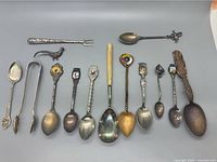 Top-down view of thirteen assorted souvenir spoons and utensils arranged horizontally on a grey background, showing various sizes, handles, and decorative elements including crests and enamel.