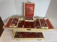 Top view of cream-colored vintage jewellery box with red interior showing multiple compartments filled with assorted vintage costume jewellery pieces including necklaces, brooches, and earrings.