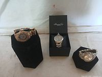 Three wristwatches including a Kenneth Cole in black box and two gold-tone watches on black display stands against white background.