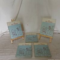 Five children's books titled The New We Three displayed on small wooden easels and laid flat on a beige surface against a white backdrop.
