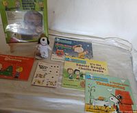 Photo showing six Charlie Brown-themed children's books and a small Snoopy plush toy arranged on a light surface. Books include holiday and character-specific themes like Christmas, Easter, Thanksgiving, and sleep time.