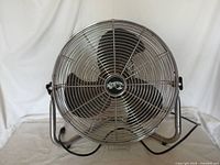 Front view of industrial fan showing chrome protective grille and three metal blades inside. Large circular shape. Power cord visible at base.