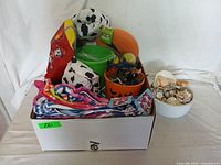 Box containing four folded colorful kids beach towels, an orange bucket, a green bucket, a netted bag with sand toys, and a red floatie with matching vest with Dalmatian pattern.