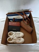 Photo showing all items in the lot inside a cardboard box: navy and white striped handbag, three clutches (pink, brown monogram, and black textured), and white sneakers.