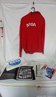Red NASA sweatshirt hanging on hook with white NASA front logo, viewed frontally with memorabilia items on table below
