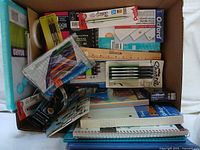 Photo showing new Sharpie permanent markers, assorted colored pens, spiral notebooks, ruled paper, photo frame kit, and a mini whiteboard in original boxes and packaging inside an open cardboard box.