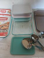 Glass Pyrex baking dishes stacked with assorted pastel lids and two stainless steel heart-shaped measuring cups with handles in front.