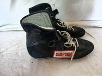 Pair of black Simpson racing shoes with white shoelaces, side view showing logo patch.