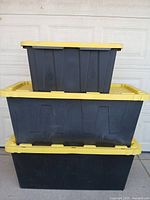 Stacked view of two large black plastic storage bins with yellow lids and one smaller bin on top. Shows dirt and marks from storage use.