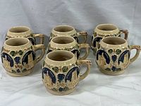 Set of six beige ceramic cups with German castle and grapevine reliefs, arranged on cloth background.