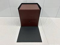 Front view of the Corum brown leather watch box with the black front flap open, showing the embossed logo and drawer.