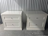 Two white 2-drawer storage cabinets side by side showing front and side views with clean white painted MDF and crystal-like knobs.