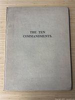 Front cover of hardcover book titled 'The Ten Commandments' in black text on beige linen cover, showing minor wear and discoloration.