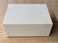 Closed view of the cream-colored rectangular cardboard Rolex outer box showing the embossed crown logo on the lid, surface has wear and scuffs.