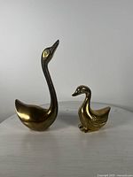 Front view of both brass duck/goose figurines showing their relative size and shape, highlighting the taller mother figurine with neck stretched upwards and the smaller baby figurine with detailed wing engravings.