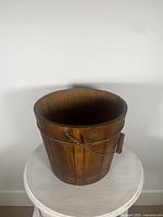 Front and side view of the antique wooden sap bucket showing its shape, wood grain, metal bands, and handle.