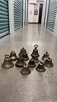Eleven antique brass bells displayed on concrete floor in storage hallway. Various sizes, dark patina, inscribed CANADIAN on bases.