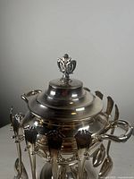 Ornate silver-plated sugar spooner bowl with lid and handle, showing decorative finial on the lid.